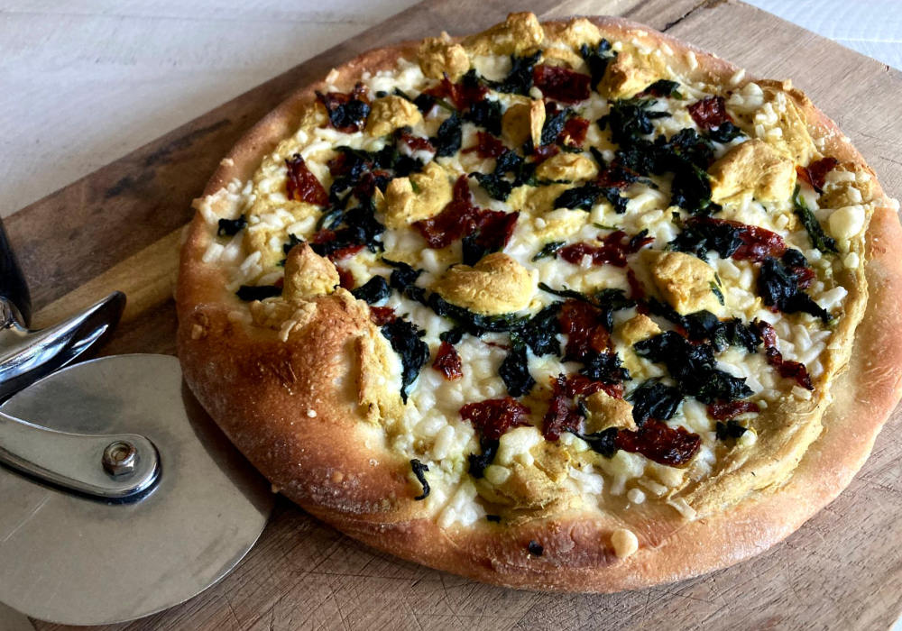 Vegan white pizza with tofu ricotta, spinach and sun-dried tomatoes unsliced on a cutting board.